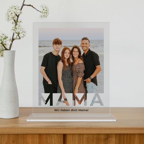 mumaboard MAMA Acrylglas Fotogeschenk mit Familienfoto am Strand und personalisiertem Spruch „Wir lieben dich Mama" neben einer Vase mit Kirschblütenzweig